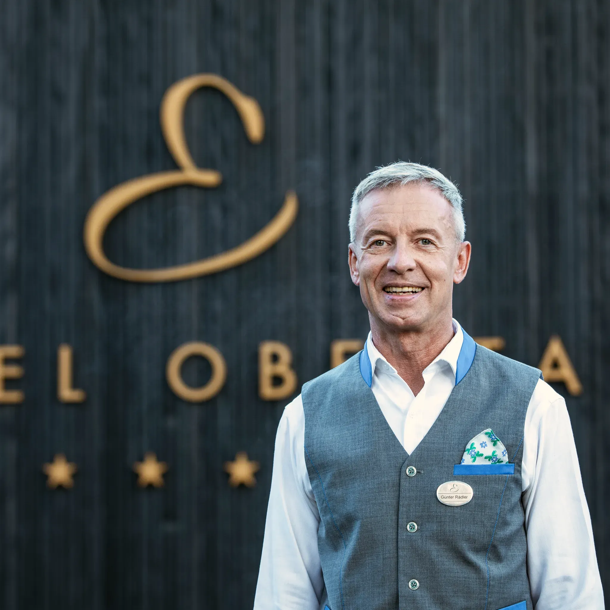 Porträt von Günter Rädler, Barchef im Engel Obertal, in Weste und Krawatte vor dunkler Holzwand mit goldenem Schriftzug.