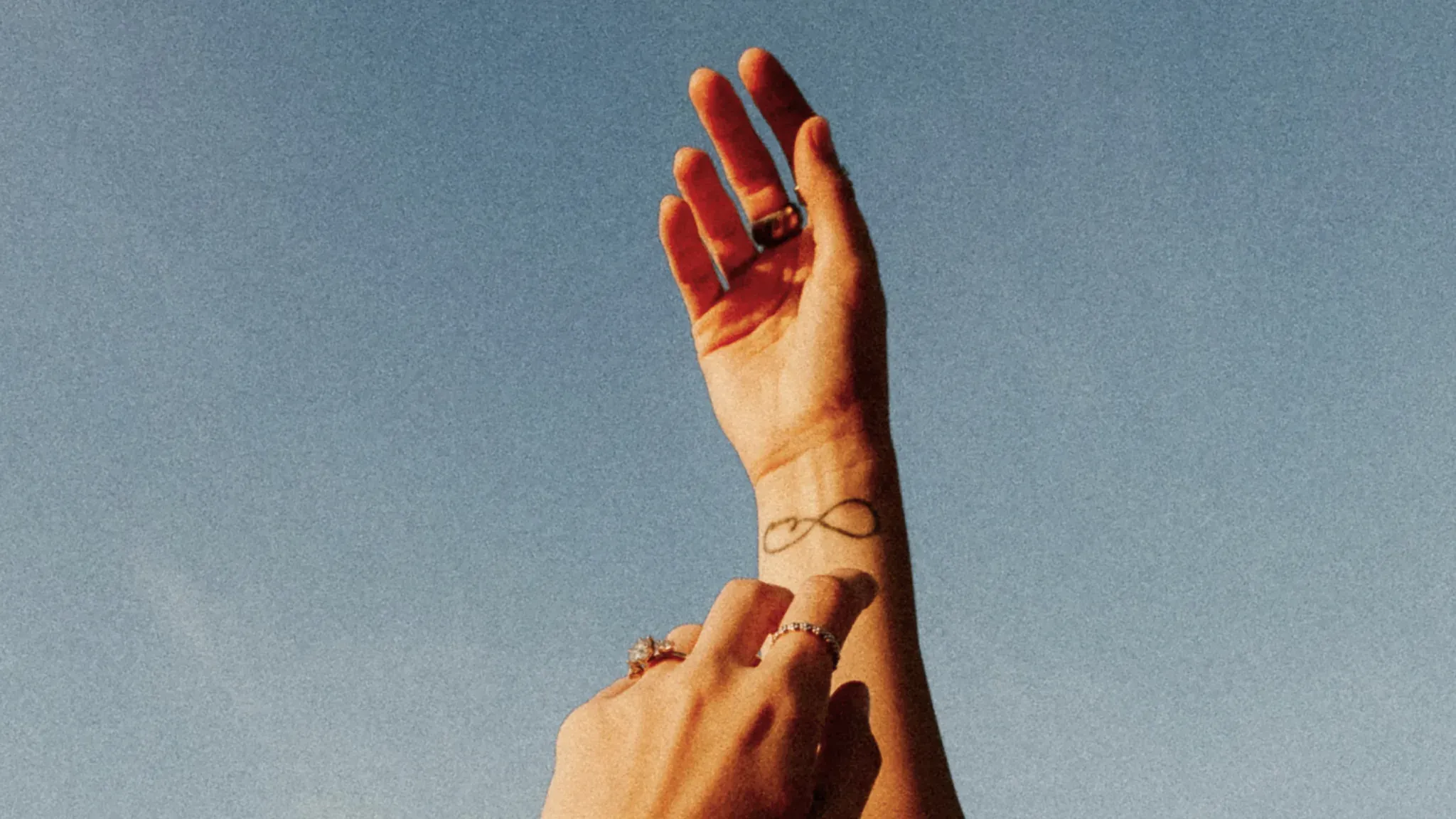 Zwei Hände berühren sich vor blauem Himmel, am Handgelenk ein Unendlichkeitstattoo.
