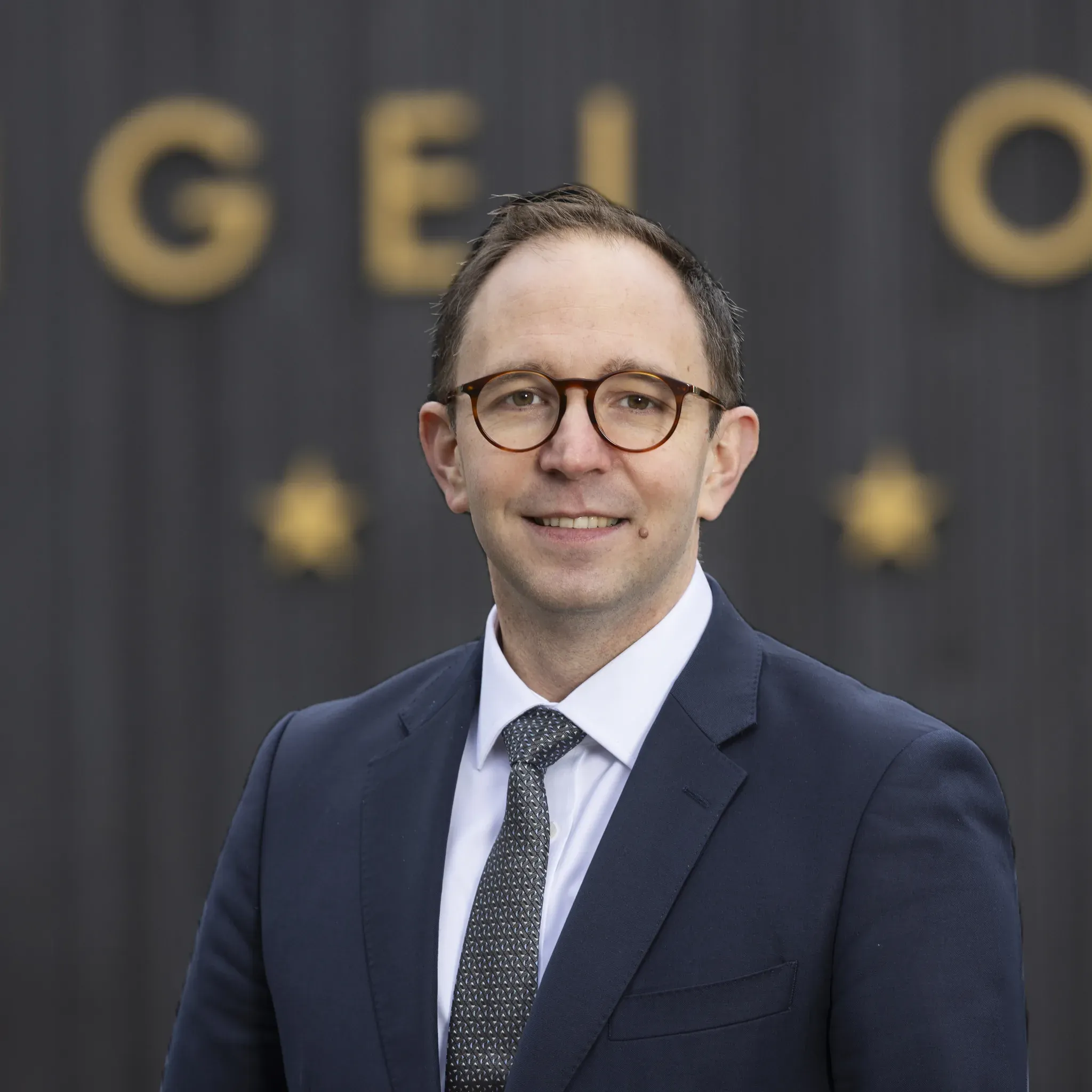 Porträt von Simon Seis, Hoteldirektor im Engel Obertal, vor dunkler Holzwand mit goldenem Schriftzug.