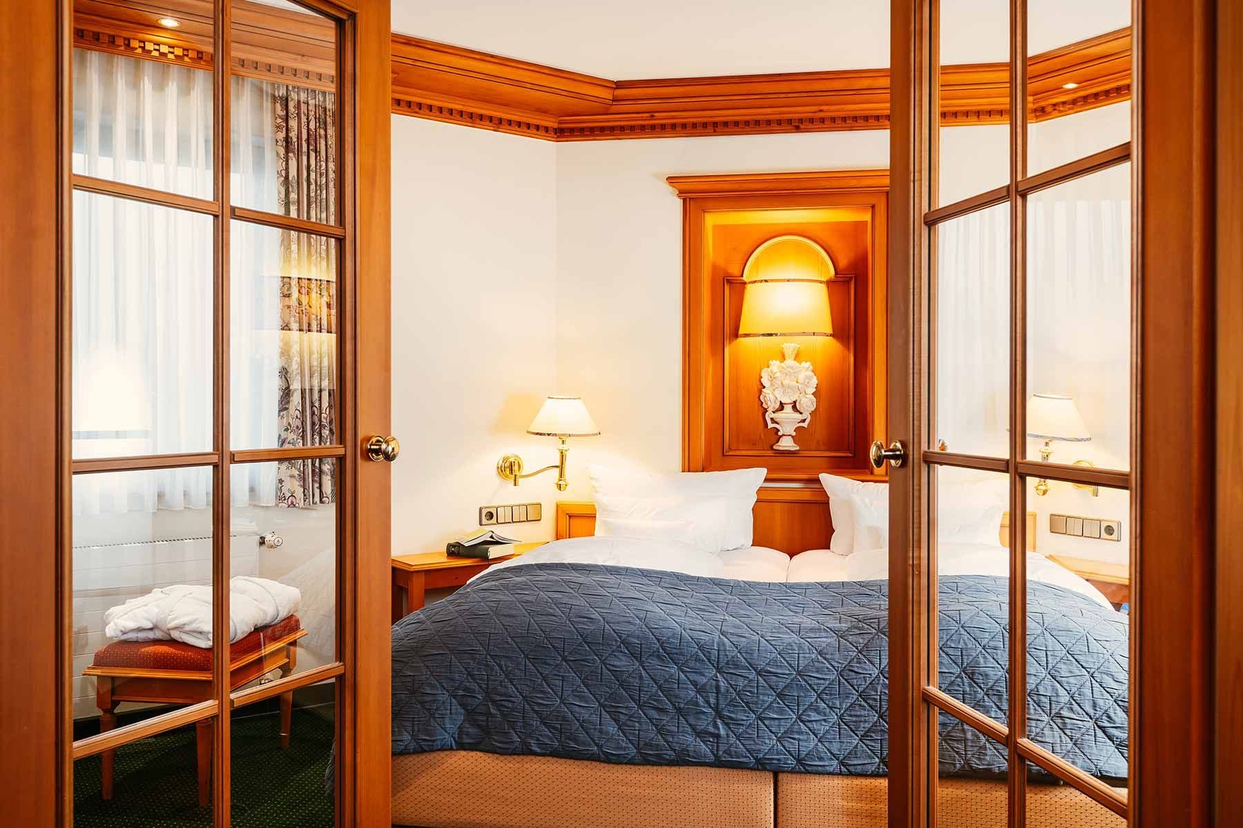Traditionelle Suite Sonnenblick im Engel Obertal – Blick durch Sprossen-Glastüren auf das Doppelbett mit blauer Tagesdecke, warme Kirschholzvertäfelung und beleuchtete Wandnische mit Blumendekor