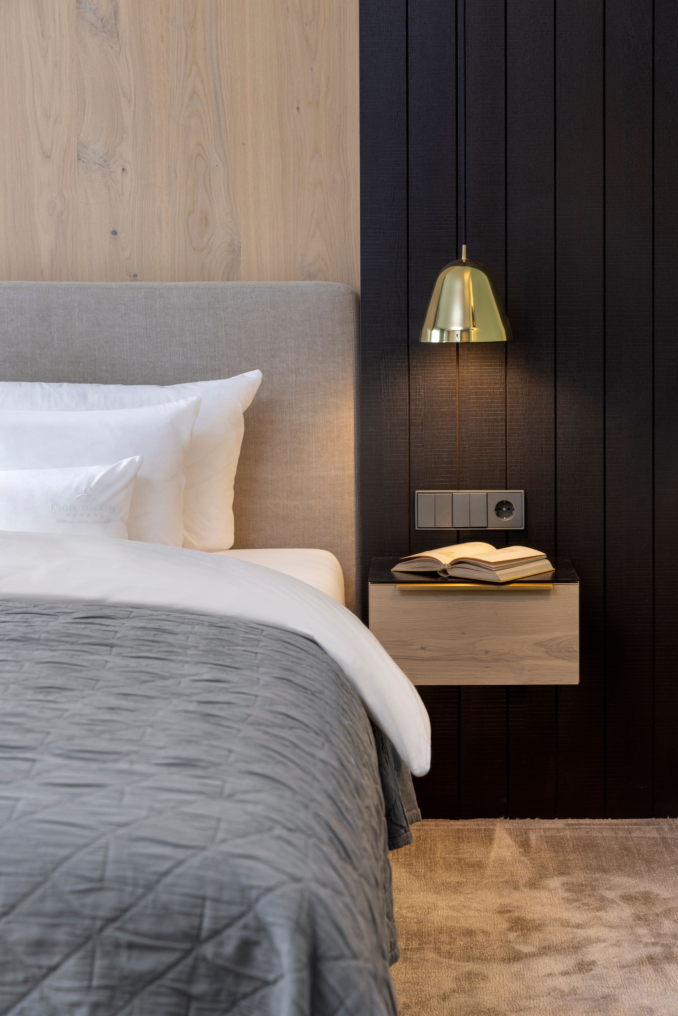 Hotelzimmer mit bezogenem Bett, dunkler Holzvertäfelung und schwebendem Nachttisch mit aufgeschlagenem Buch unter einer Messing-Wandleuchte.