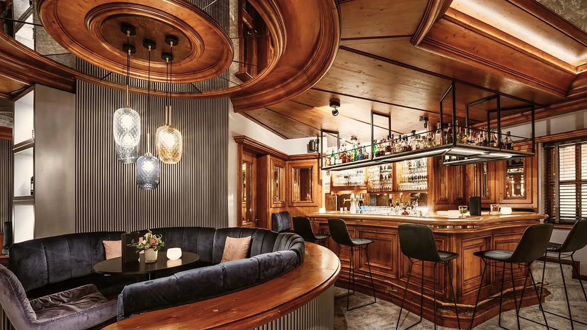 Bar mit heller Holzvertäfelung, geschwungener Polsterbank, Barhockern und Flaschenregal.