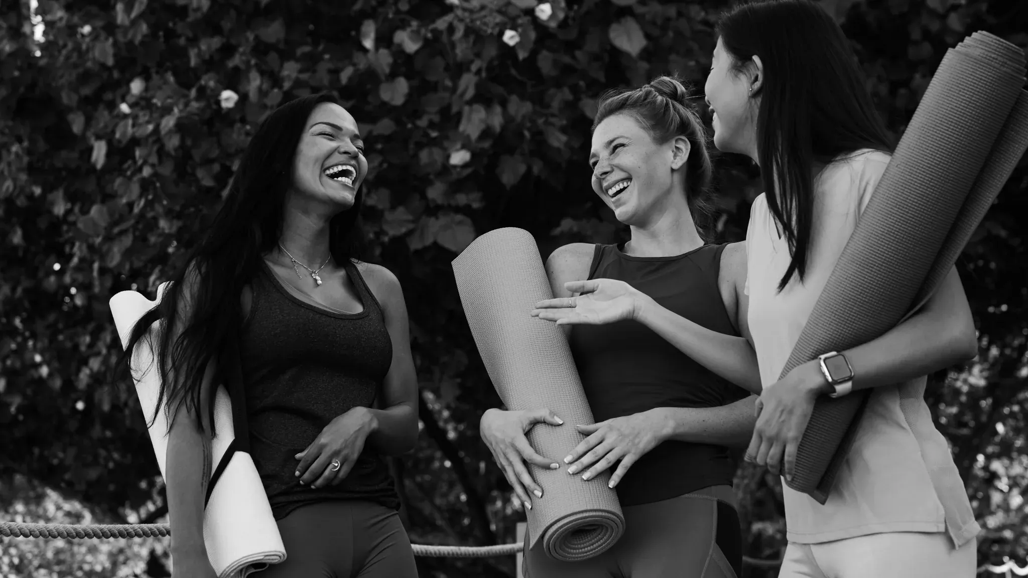 Drei lachende Frauen mit Yogamatten unter dem Arm im Gespräch, Schwarzweiß.