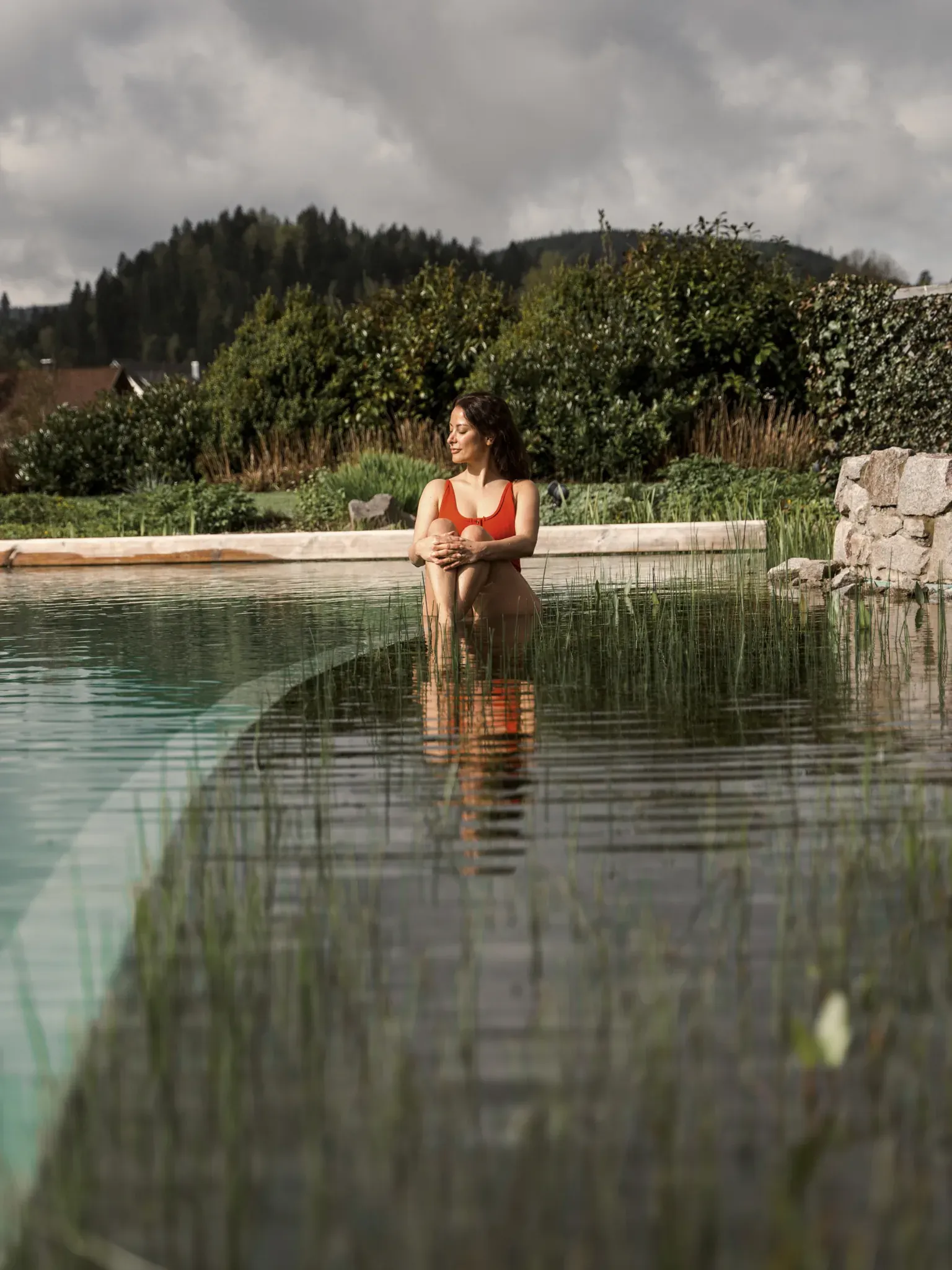 Frau in rotem Bikini sitzt im Wasser am Rand eines Naturbadeteichs vor bewaldeten Hügeln.