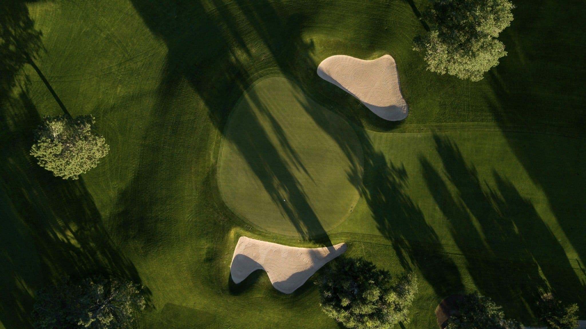 Luftaufnahme eines Golfplatz-Grüns mit zwei hellen Sandbunkern, Bäumen und langen Schatten.