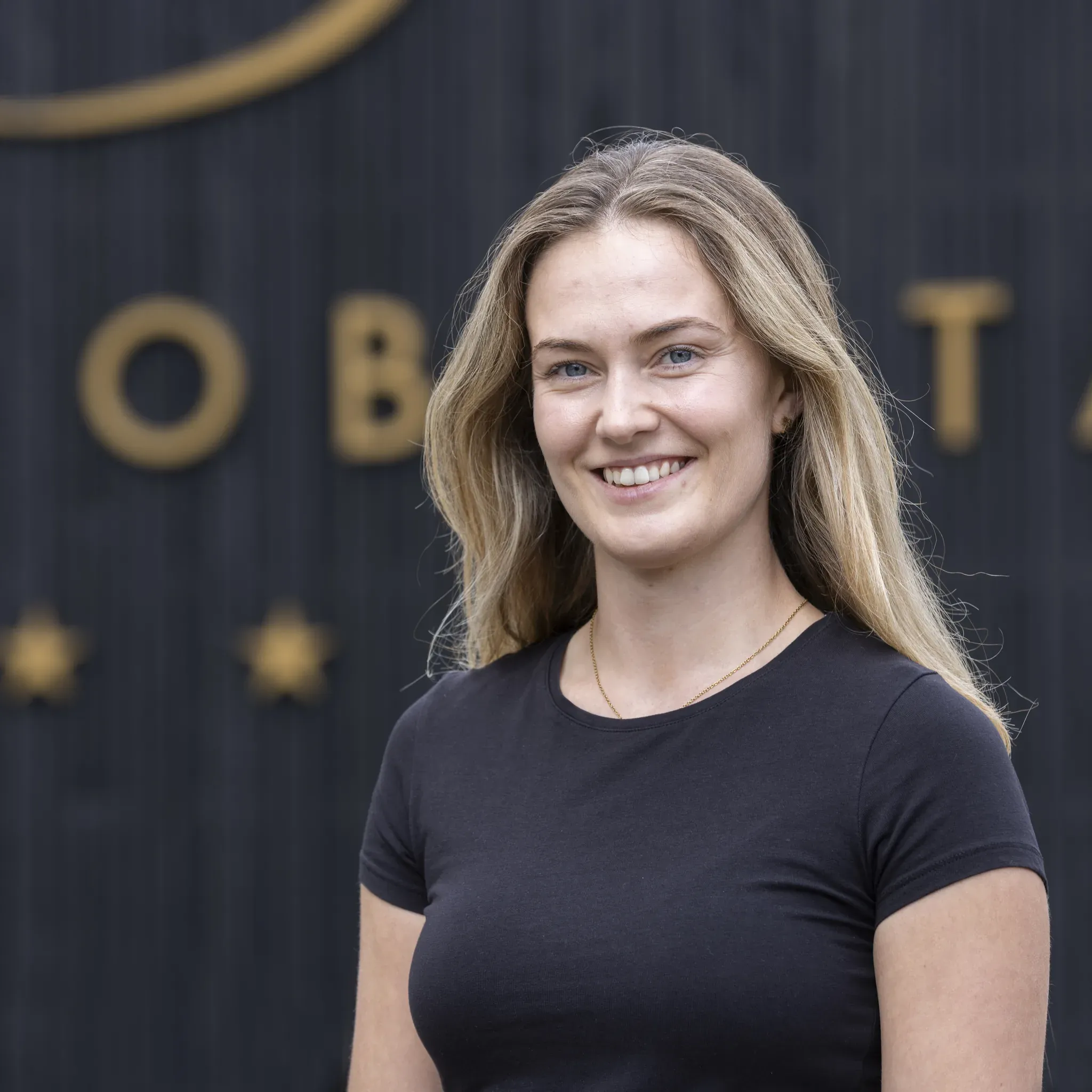 Porträt von Jessica Braun, zuständig für Sport und Gästeunterhaltung im Engel Obertal, vor dunkler Holzwand mit goldenem Schriftzug.