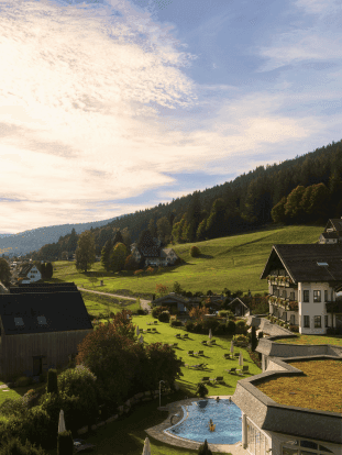 Luftaufnahme der Hotelanlage mit Chalets, Wiesen und einem Außenpool vor bewaldeten Schwarzwaldhügeln.
