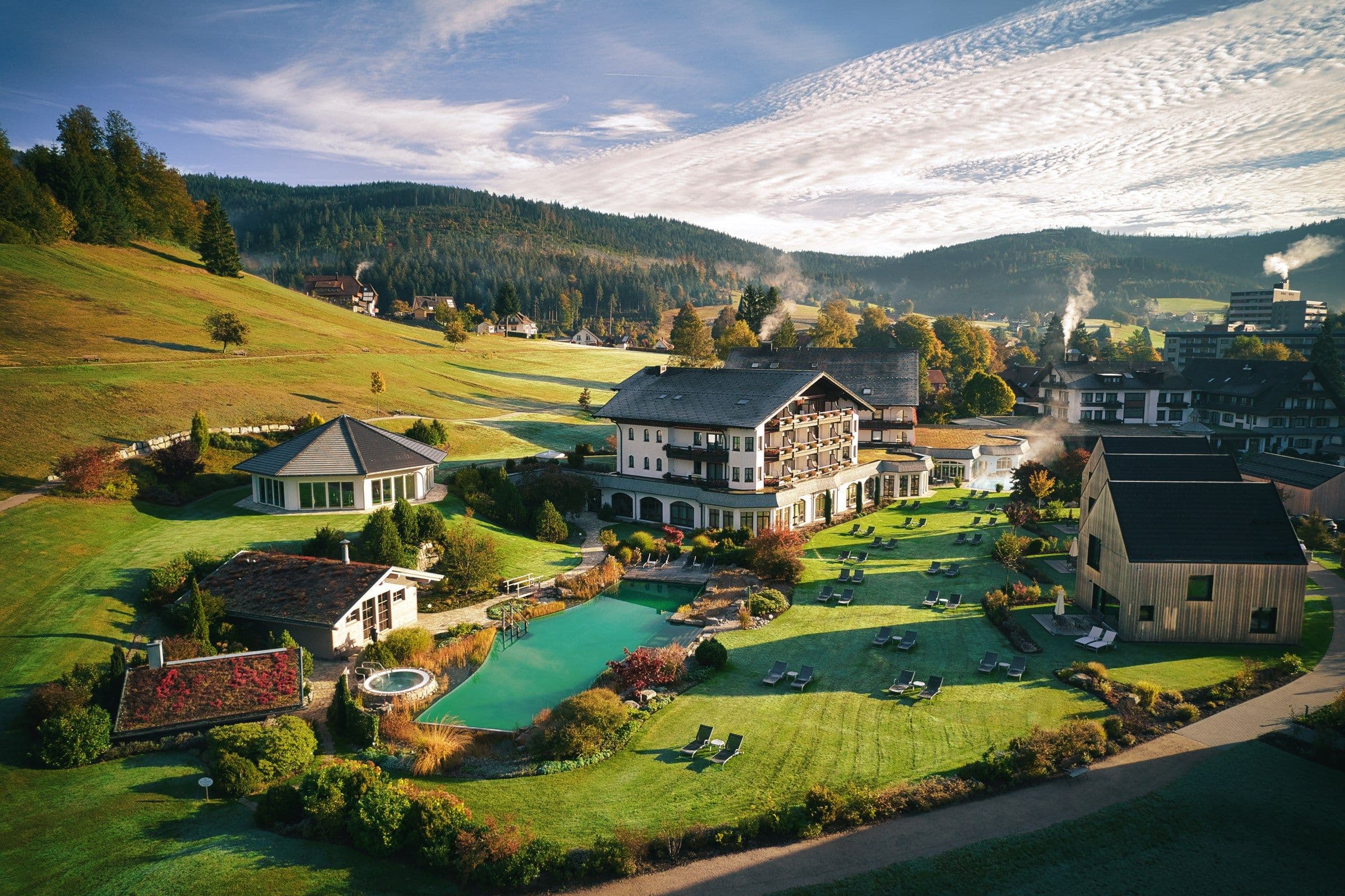 Luftaufnahme des Engel Obertal: Hotelgebäude, Chalets, Naturbadesee und Liegewiese im Talkessel, umgeben von Schwarzwaldhängen im Herbstlicht.