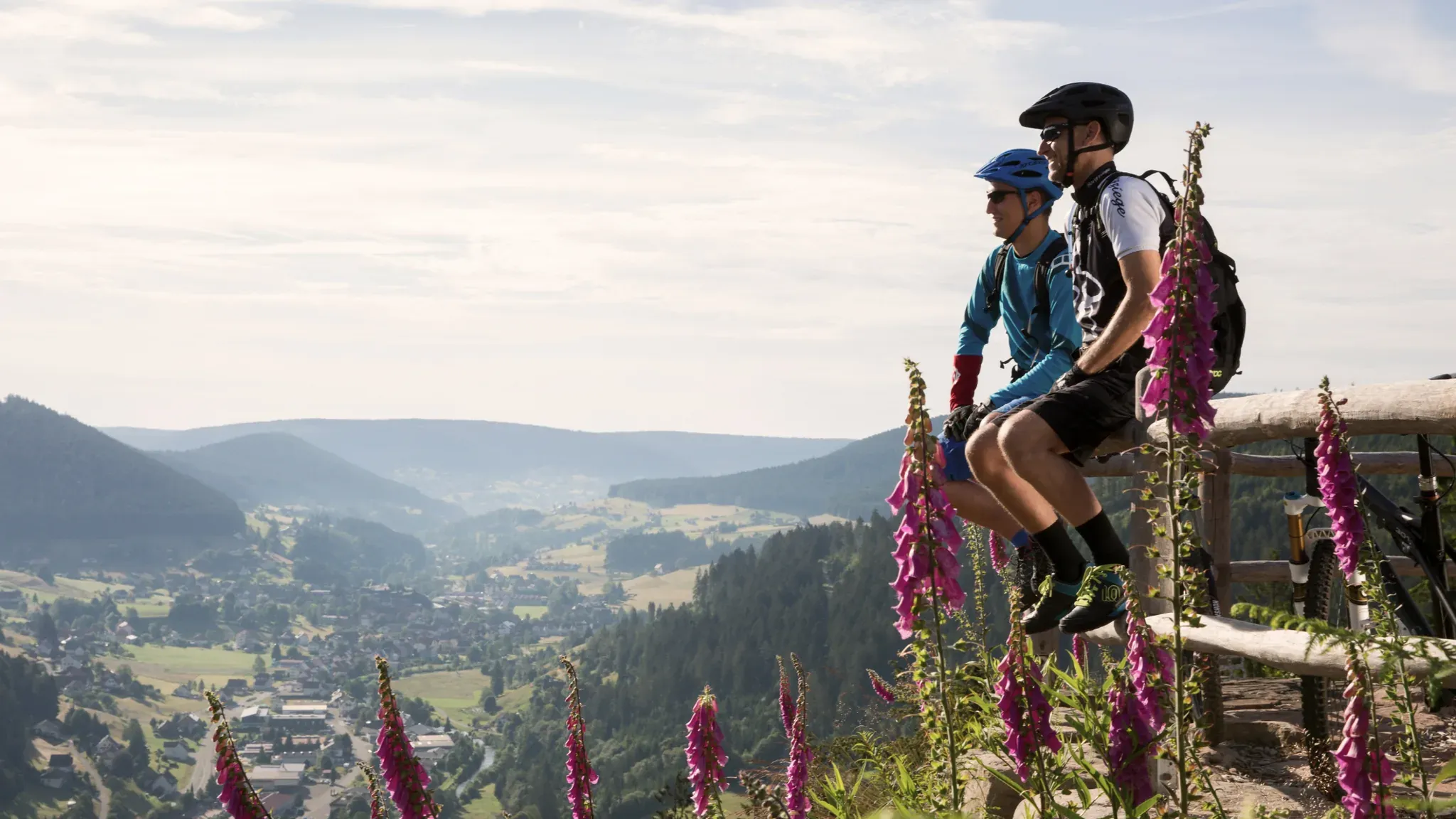 Zwei Mountainbiker sitzen auf einer Bank über dem Tal, im Vordergrund blühender Fingerhut.