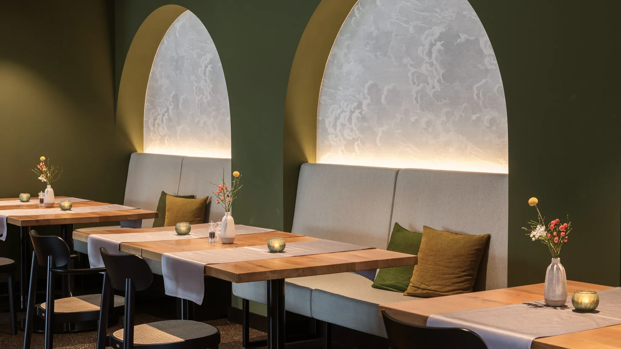 Restaurantbereich mit olivgrüner Wand, hinterleuchteten Bogennischen und hellen Holztischen.