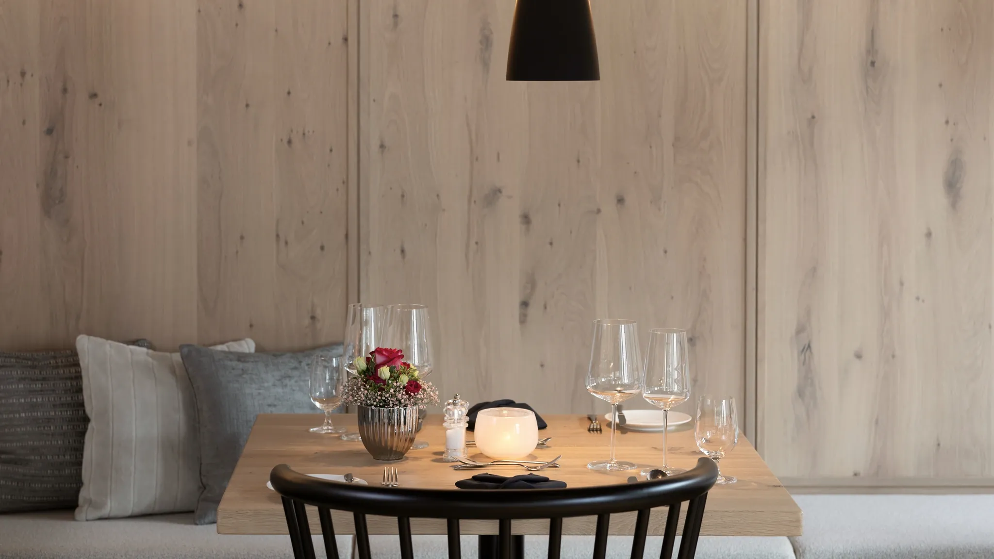 Runder Tisch mit Weingläsern, Blumen und Teelicht an einer hellen Holzwand unter einer schwarzen Pendelleuchte.
