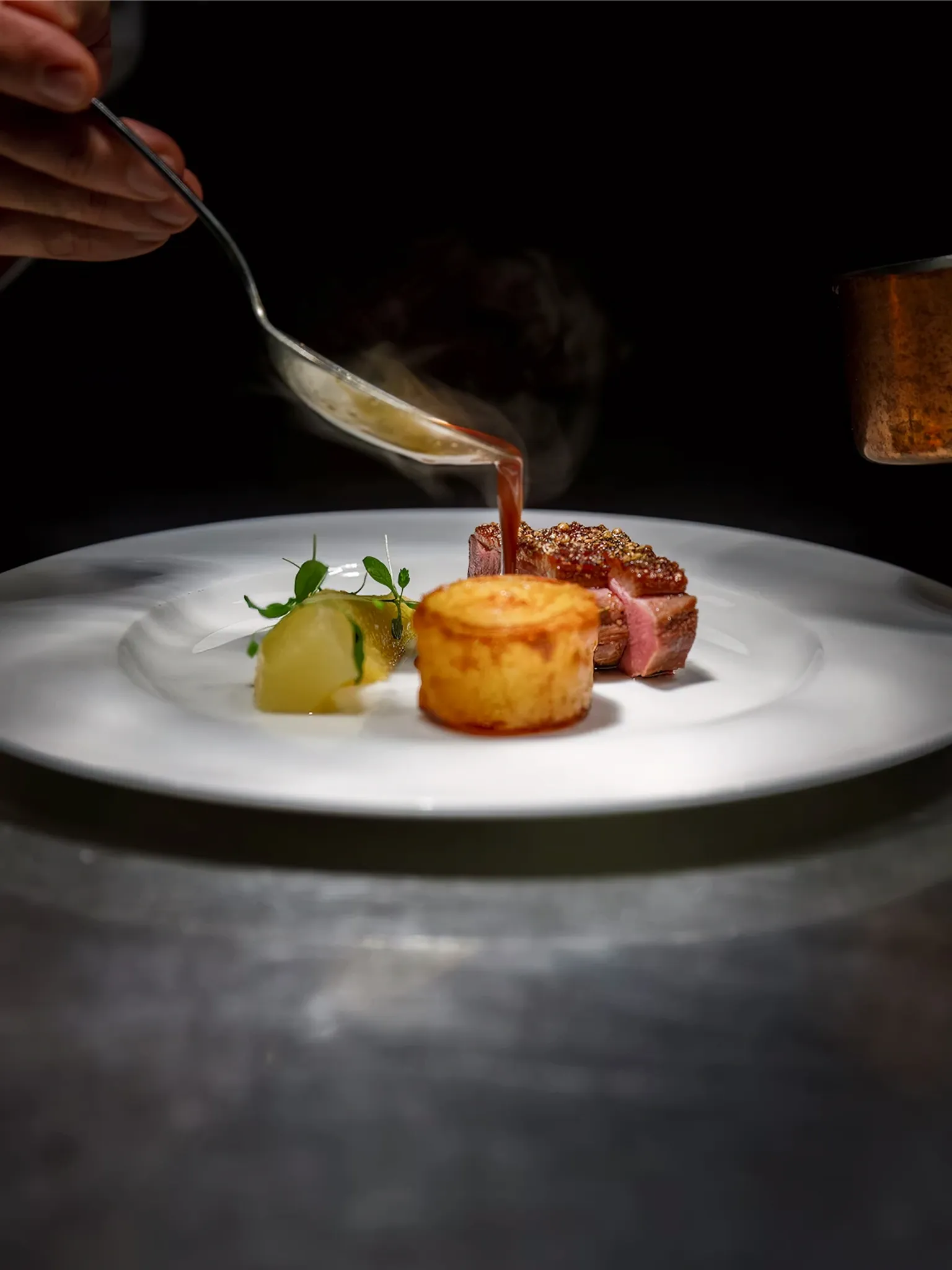 Aus einer kleinen Kanne wird dunkle Sauce über ein angerichtetes Gericht auf weißem Teller gegossen, dunkler Hintergrund.