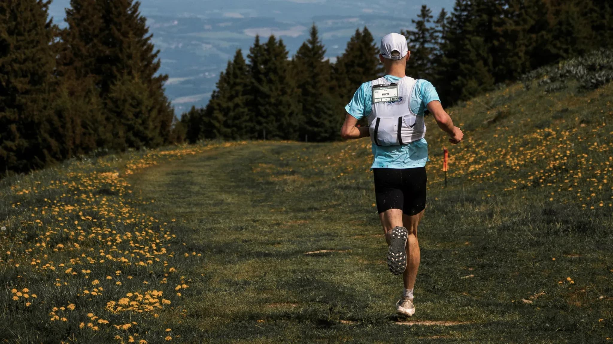 Läufer mit Trinkweste joggt über eine blühende Bergwiese am Waldrand.