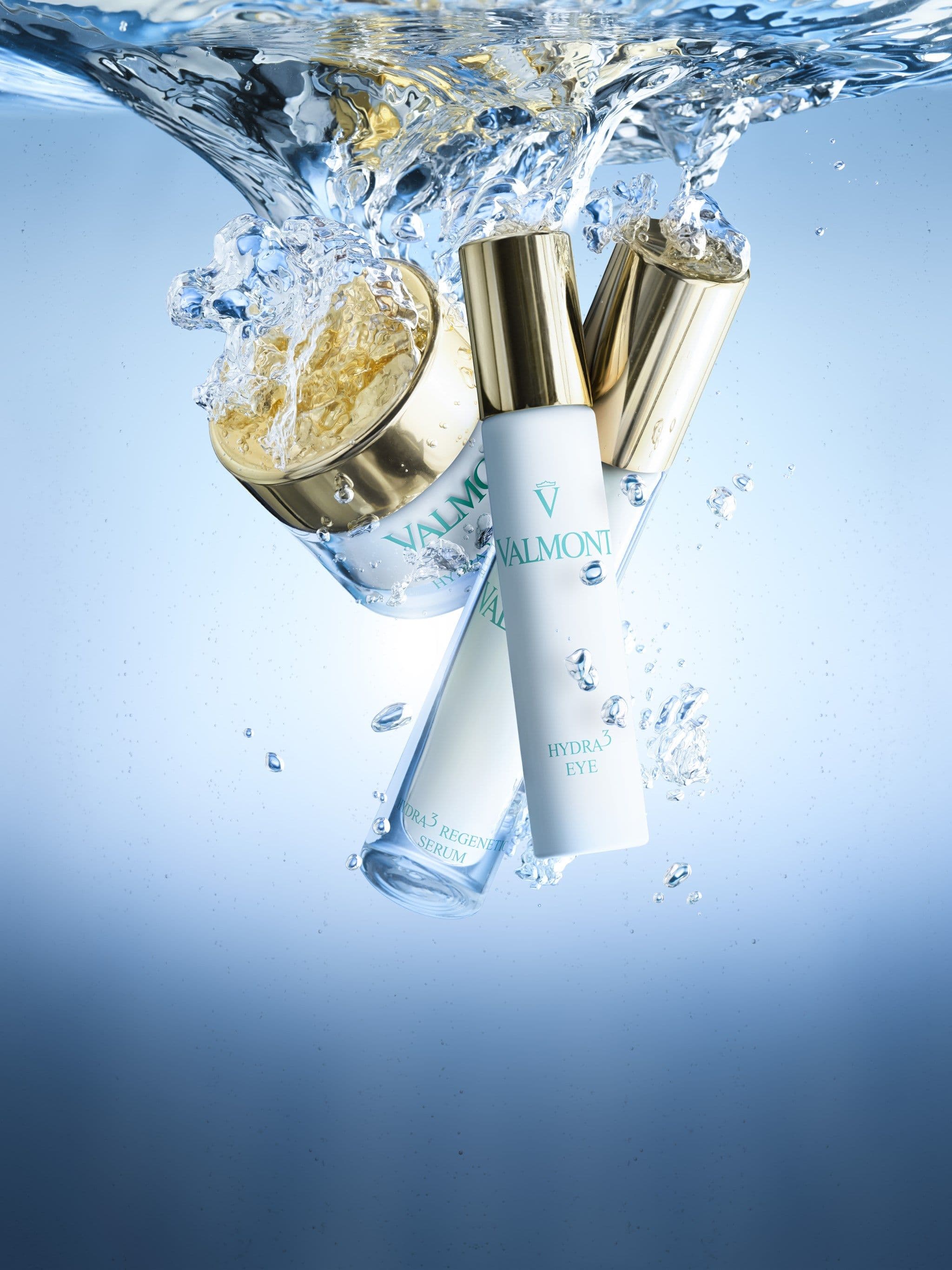Drei Pflegeprodukte der Linie Maison Valmont – Cremetiegel, Regenerating Serum und Hydra3 Eye – mit goldenen Verschlüssen tauchen in klares blaues Wasser ein.