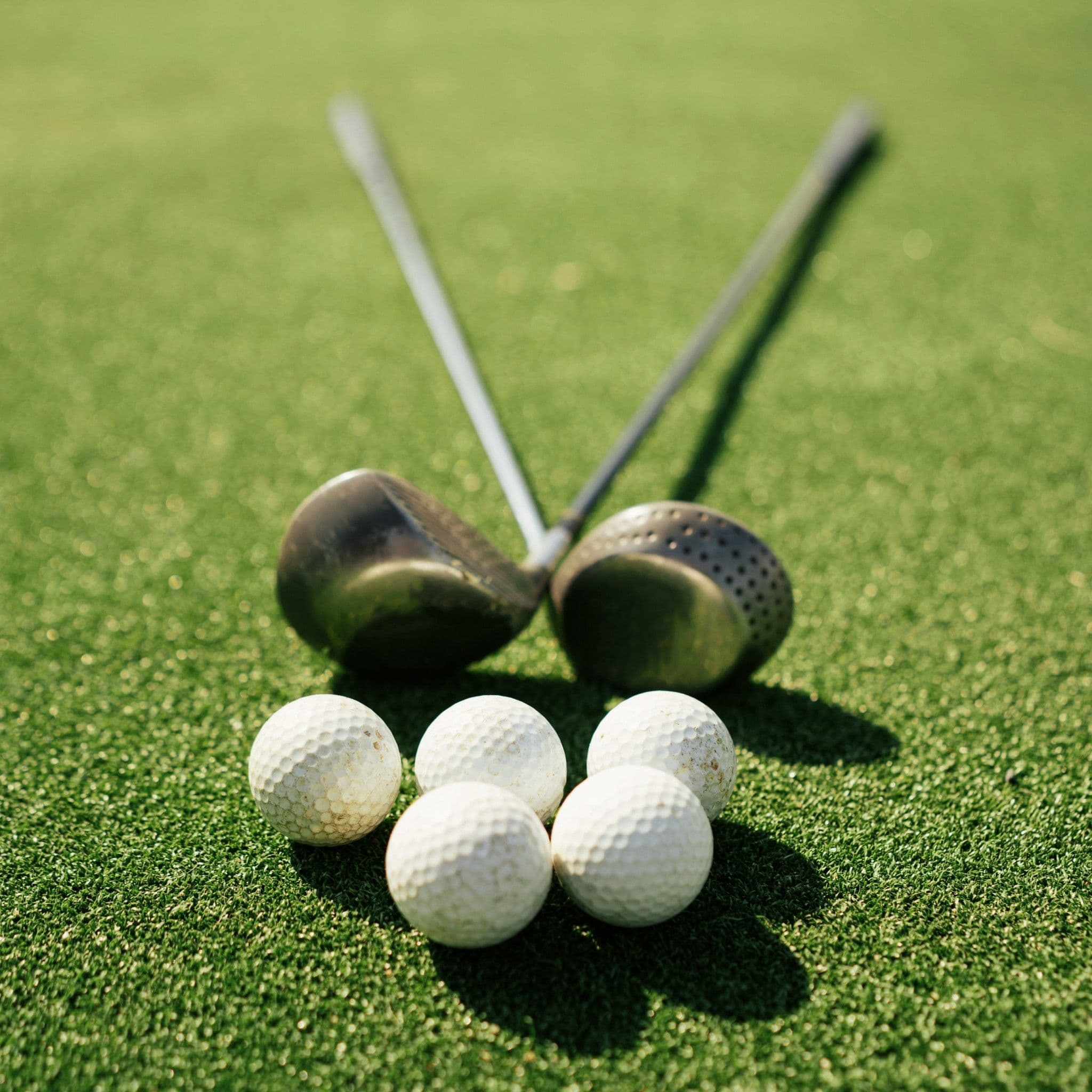 Die Köpfe zweier gekreuzter Golfschläger hinter fünf Golfbällen auf sonnigem Rasen.