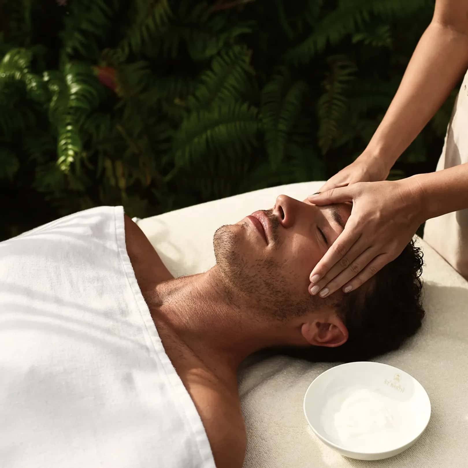Mann liegt mit geschlossenen Augen auf einer Behandlungsliege, während eine Person sanft sein Gesicht massiert. Neben der Liege steht eine Schale mit weißer Creme.