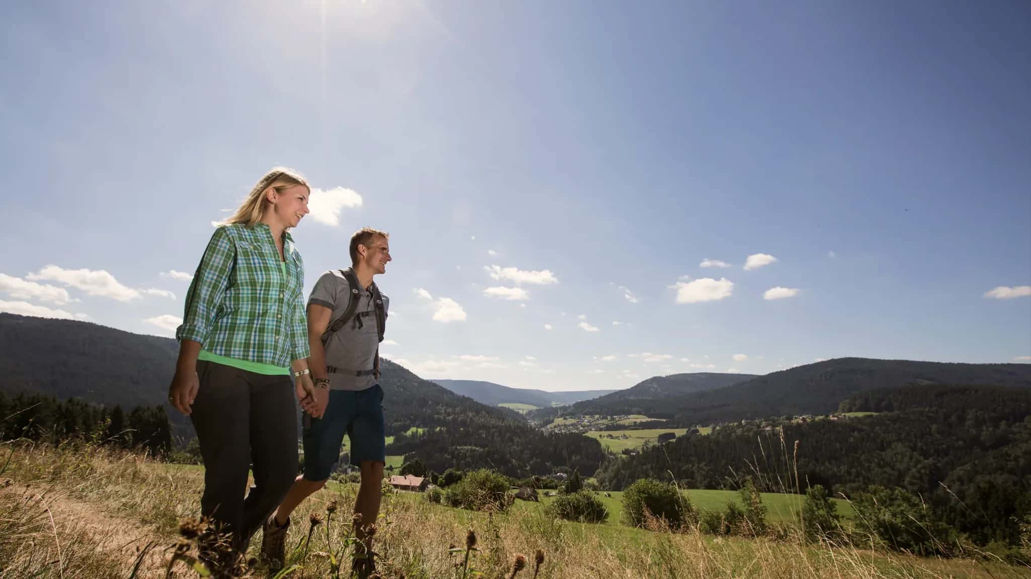 Ein Paar wandert über eine Höhenwiese mit weitem Blick über die Schwarzwaldberge.
